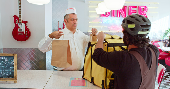 A man in a white button down shirt puts an order inside a delivery person's bag in a 1950s styled diner.