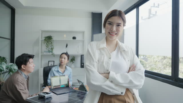 Waist-up view of cheerful corporate asian woman professional with arms crossed looking at camera, while associates converse in background.