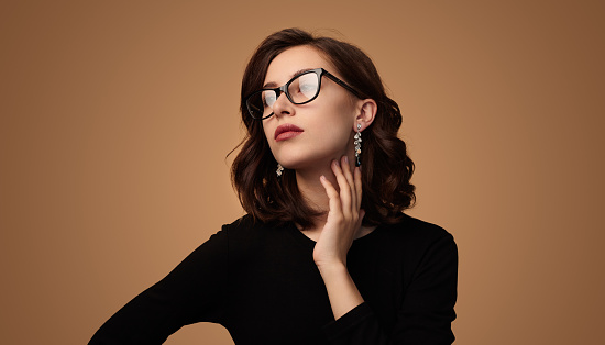 Stylish young brunette with elegant glasses and earrings touching neck and looking at camera against brown background