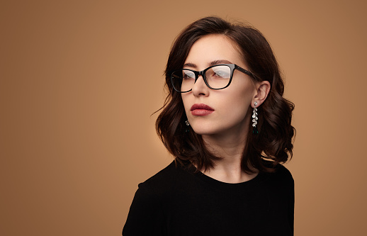Confident young female model with dark wavy hair and makeup in elegant clothes and eyeglasses looking away against beige background