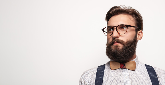 Clever bearded man in stylish clothes and glasses looking away with serious face expression against gray background
