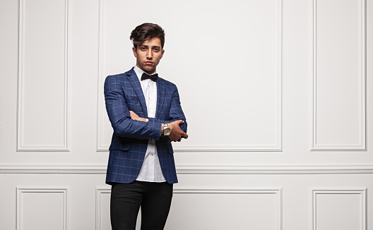 Self assured young male in smart casual clothes crossing arms and looking at camera while standing against white wall