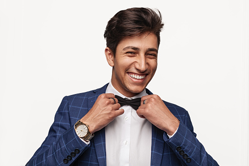 Happy young ethnic man in elegant suit with luxury wristwatch looking at camera with smile and adjusting bow tie against gray background