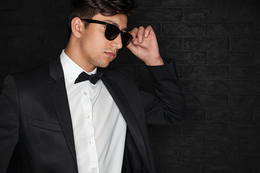 Young ethnic male in elegant suit adjusting stylish sunglasses and looking away against black brick wall
