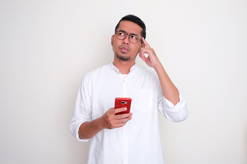 Adult Asian man thinking about something while holding a mobile phone