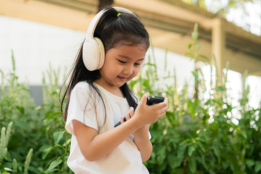 Toddler girl wearing headphone listen music on mobile phone at park. experience of using devices and positive expressions