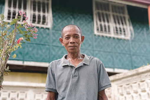 A simple tanned southeast asian man in his early 60s posing in front of his rural two storey house.