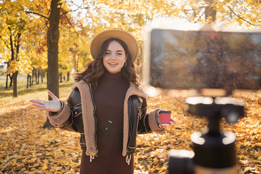 Female blogger recording video outdoors. Beautiful young girl smiling and looking at the camera standing in the autumn park, talking and gesticulating with both hands. Blogging, video blog and people
