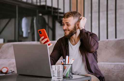 Puzzled caucasian beardy caucasian man in casual sitting on couch at desk with laptop holds phone looks at screen hesitates. Pensive European guy remote works home. Troubles, perplexing male.