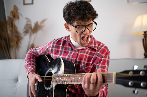 Young arabic man playing acoustic guitar and screaming song sitting at home. Copy space