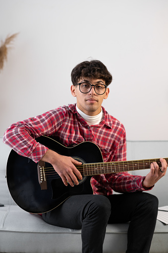 Handsome young man in casual clothes is looking at camera while playing guitar at home. Copy space