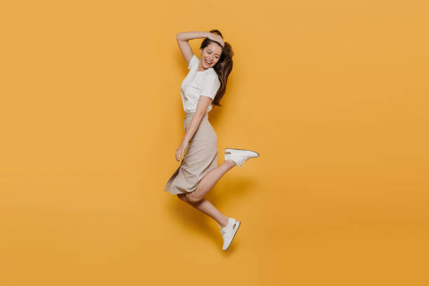 Cheerful pretty brunette in white t-shirt, beige skirt and white sneakers, jumping high, careless mood, enjoying her life, ready for summertime. Studio shot over yellow backdrop. Happy people concept. Cheerful pretty brunette in white t-shirt, beige skirt and white sneakers, jumping high, careless mood, enjoying her life, ready for summertime. Studio shot over yellow backdrop. Happy people concept. jump jet stock pictures, royalty-free photos & images