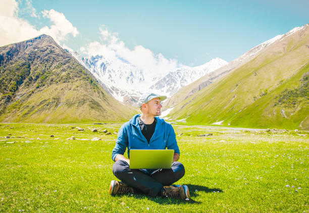 Caucasian handsome guy digital nomad sit work type on laptop with mountains background. Digital nomad traveler blogging on travels. Ideas and work online in post pandemic world.