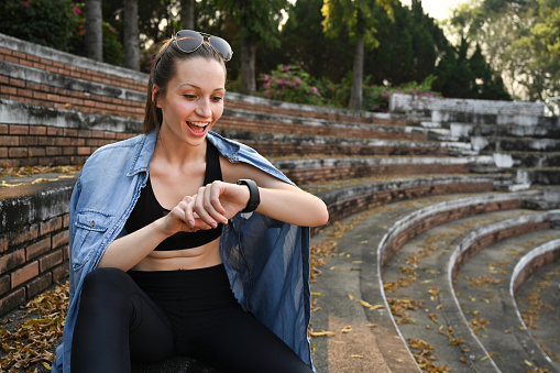 Happy athletic woman checking fitness results or heart rate on smartwatch after running in the park.