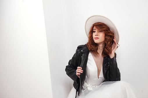Happy stylish bride pretty lady in white wedding dress, hat, leather jacket posing relaxing, looking away. Fashionable young german cute woman poses. Fashion style beauty concept. Copy ad text space