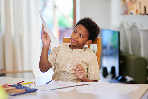 Homeschool boy child, paper plane and playing at desk in home living room for learning, education or task. African kid, play and studying in house, apartment or room for creative development at table