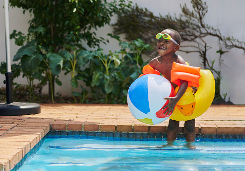 Swimming, black child playing in pool with ball, smile and fun time in water with toys in backyard in summer. Swim, play and sun, boy with beachball and happy, safe activity for young kids on weekend