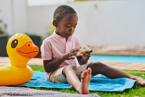 Food, lunch and black kid with sandwich at backyard outdoors by swimming pool. Nutrition, health and hungry, sad and young black child or boy sitting on towel and eating meal for wellness alone.