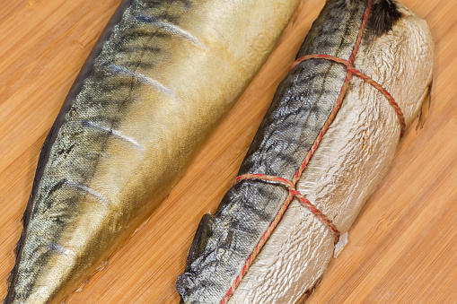 One hot-smoked Atlantic mackerel tied with twine and one cold-smoked mackerel without heads on a wooden cutting board, fragment close-up