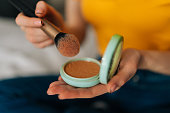 Close-up hands of unrecognizable young woman holding make up powder brush and case using it sitting on bed in bedroom. Concept steps of make-up applying.