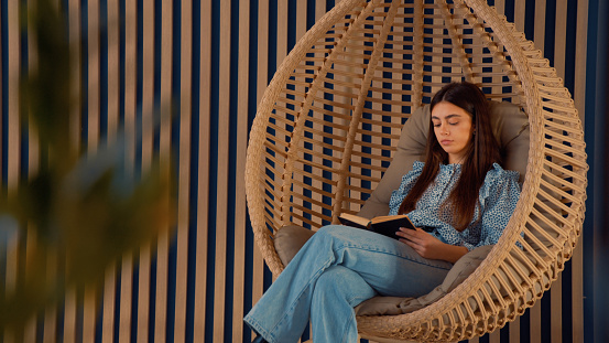 Young woman is relaxing and reading a book sitting in a cozy hanging  swing chair with cushions