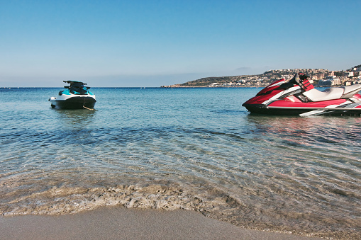 A couple of recreational power jet skis with no people at the water's edge of a sandy beach shoreline in the Mediterranean sea