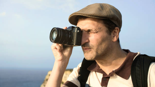 istock photographer is taking photo outdoors with mirrorless camera with blue sky background with copy space. Photographer man with modern camera outdoors taking photos. close up view 1476130290