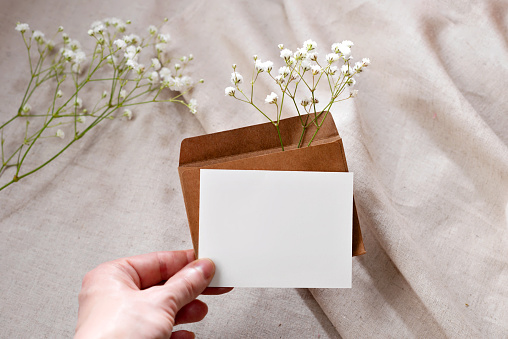 Blank paper card and envelope with flovers in persons hand on a beige background, elegant invitation, greeting card or business card template with mockup copy space