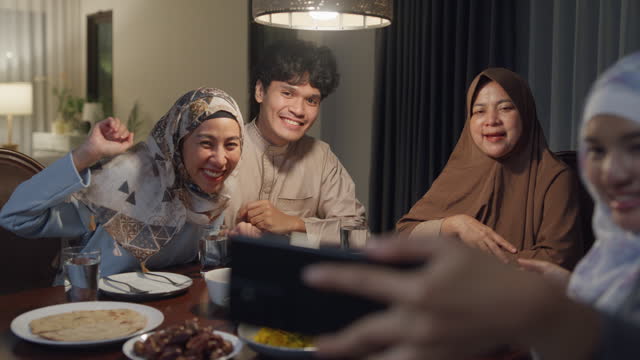 Happy Asian muslim family celebrate long distance with cousin use cell phone video call online Ramadan dinner together at home. Two generation celebration of Eid al-Fitr togetherness.