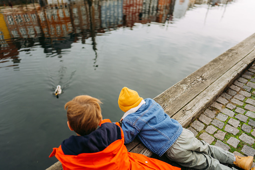 Photo is a reflection in water of colourful traditional houses in Nyhavn pier, Copenhagen,  Denmark. Two boys are bent over the Canal and feeding the duck