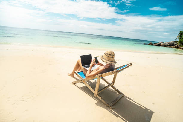 istock Photo fo a woman in summer hat using mobile phone and laptop computer, working while sitting on a beautiful tropical beach. Digital nomad. 1476151389