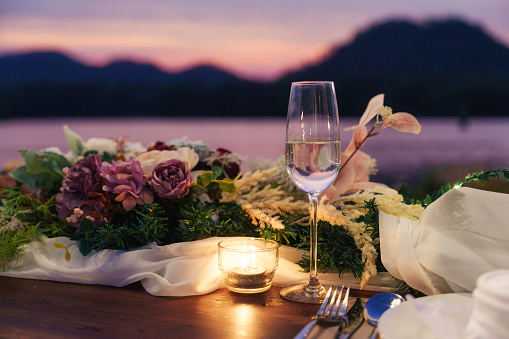 Rustic style wooden dining long table with flower, wine glass, candle decoration at dinner by the river in the sunset