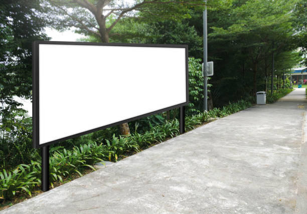 istock Large horizontal blank advertising poster billboard mockup, framed by trees and plants, lush surrounding. Digital light box display screen for OOH media. Out of home template beside pedestrian path 1476161728