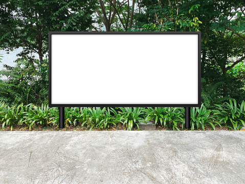 Large horizontal blank advertising poster billboard mockup, framed by trees and plants, lush. Straight digital light box display screen for OOH media. Out of home template beside pedestrian path.