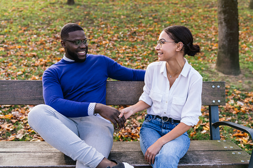 A heartwarming portrait of a black teenage boy smiling while sitting with a happy caucasian girl on a park bench surrounded by fallen leaves, basking in the warm glow of a beautiful sunset.