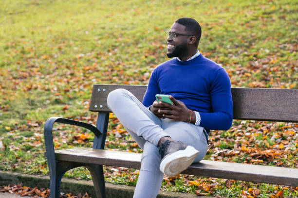 istock oung African man, casually dressed in blue sweater and glasses, happily chatting on phone while sitting on park bench in great lighting 1476172331