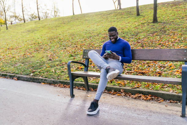 Young African man in casual blue sweater and glasses chatting on mobile phone, seated on park bench with great lighting