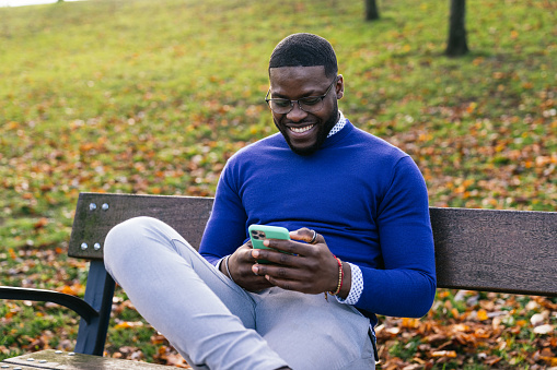 A young African man sits on a park bench, dressed casually in a blue sweater and wearing glasses. With a wide smile on his face, he chats on his phone, enjoying the beautiful lighting of the park.