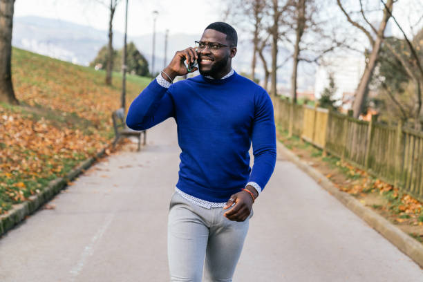 istock Young African man, casually dressed in blue sweater and glasses, beaming while talking on his phone in a well-lit park. 1476172419