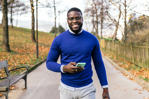 A handsome young African man is posing in a park, dressed casually in a blue sweater and glasses. He's captured with a big smile on his face, holding his mobile phone in hand, enjoying the beautiful natural light in the background.