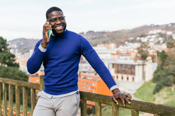 Young handsome African man in casual blue sweater talking on phone, overlooking city from park railing with stunning lighting