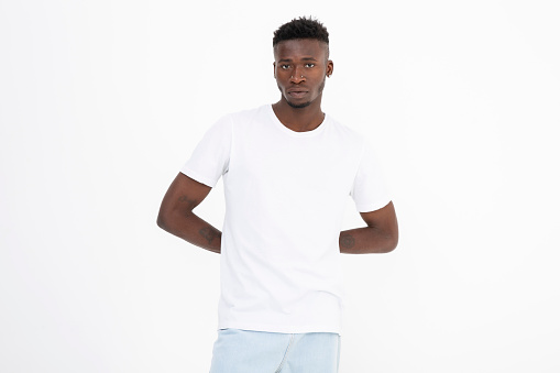 Black man of African descent wearing a white t-shirt. Expressionless. isolated on white. Hands on top of each other. Arms behind.