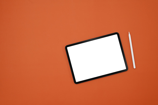 Top view with empty white screen of digital tablet, tablet and stylus pen on orange studio background.