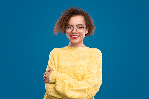 Positive female teenager with curly hair in yellow knitted pullover and glasses looking at camera against blue background