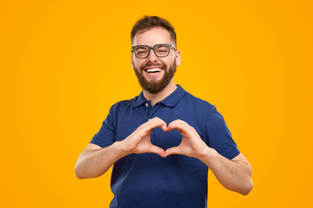 Cheerful bearded man showing heart gesture Positive adult bearded mal volunteer in eyeglasses making heart shape with fingers as symbol of peace and love against yellow background man showing peace sign stock pictures, royalty-free photos & images