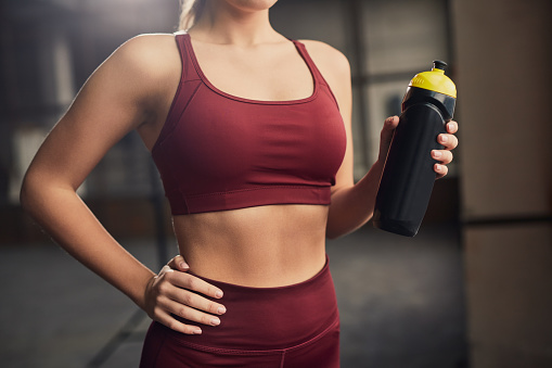 Crop unrecognizable slim female athlete in sportswear with hand on hip and bottle of water in gym