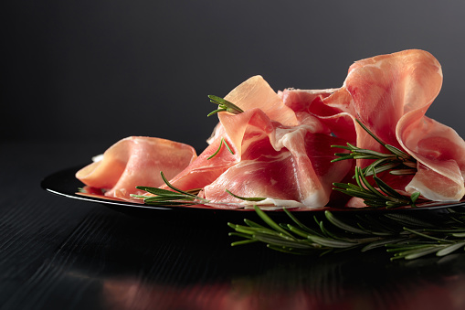 Italian prosciutto or Spanish jamon with rosemary on a black plate. Copy space.