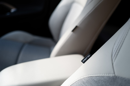 Shallow focus of an SRS Airbag tag seen attached to front part leather seats in a hybrid, new technology vehicle. The central armrest can also be seen.