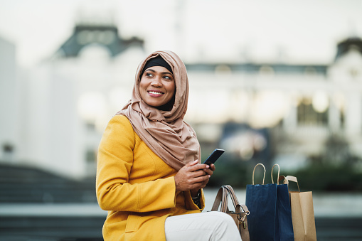 Black Muslim woman wearing hijab with a happy face sitting in urban environment and tipping messages on her smartphone.