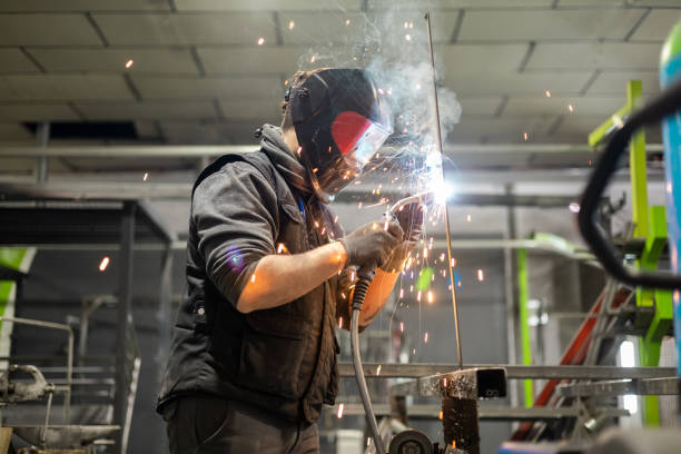 Profile view of a man working as a welder with a protective mask and work clothes performs a job with a MIG-MAG welding machine. Production in factory of welding works.